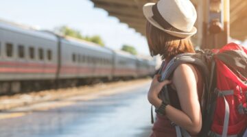 Eine junge Frau mit rotem Rucksack und Strohhut steht auf einem Bahnsteig und blickt einem einfahren