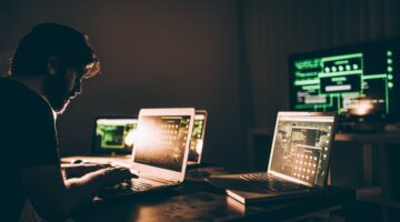 Ein Mann mit Bart und Brille arbeitet in einem abgedunkelten Raum an mehreren Laptops und Monitoren,