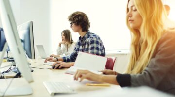 Drei junge Studierende arbeiten konzentriert in einem hellen Computerraum an Desktop-PCs, eine Stude