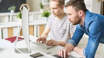 Zwei junge Männer arbeiten konzentriert nebeneinander an einem Desktop-Computer und einem Laptop in 