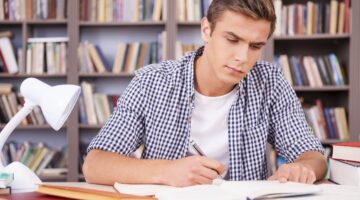 Ein junger Mann in kariertem Hemd sitzt konzentriert an einem Bibliothekstisch und schreibt mit eine