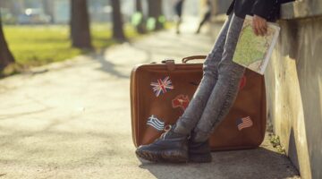 Detailaufnahme: Beine in Jeans und Wanderschuhen neben Vintage-Lederkoffer mit Laender-Aufklebern (U