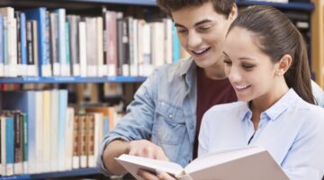 Ein junges Pärchen steht lächelnd in einer Bibliothek und schaut gemeinsam in ein aufgeschlagenes Bu