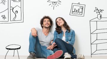 Ein junges Pärchen sitzt lächelnd auf dem Boden einer leeren Wohnung und träumt von der Einrichtung