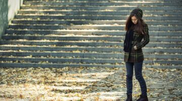 Eine junge Frau in kariertem Herbstmantel, Mütze und schwarzem Schal steht nachdenklich und den Blic