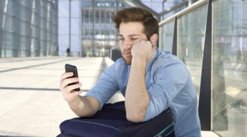 Ein junger Mann sitzt gelangweilt auf seinem Koffer an einem modernen Flughafen oder Bahnhof und sch