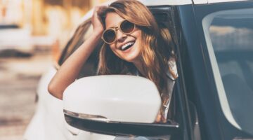 Eine lachende junge Frau mit Sonnenbrille lehnt aus dem Fenster eines weißen Autos und genießt sicht